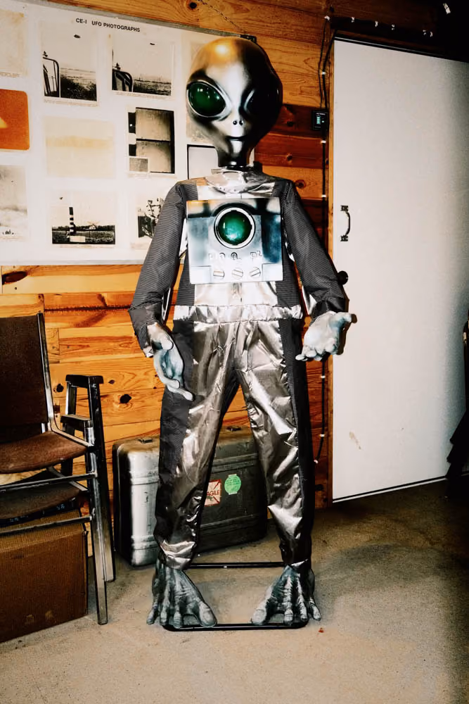 Life-size alien figure in metallic suit with large green eyes standing indoors against wood paneled wall.