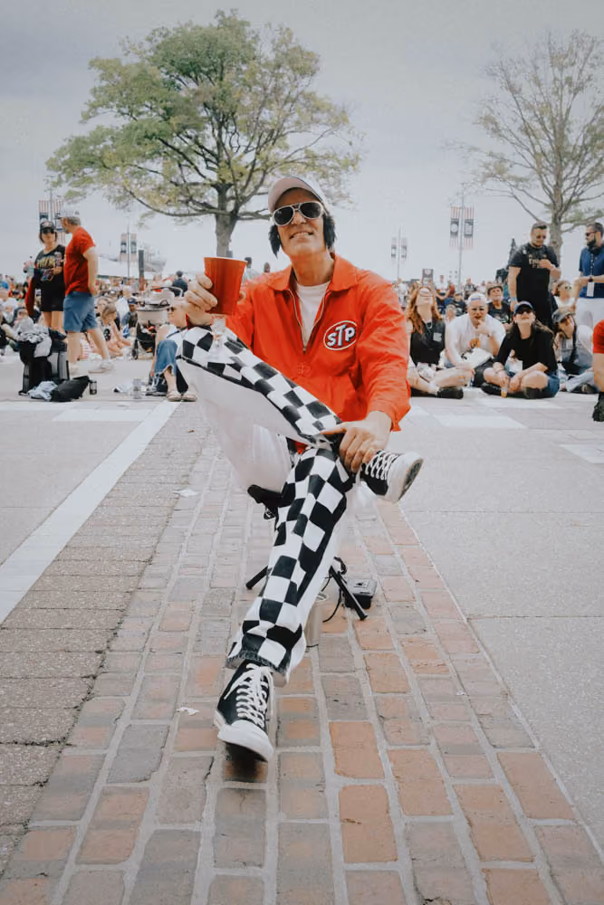 Man in checkered pants and red jacket holding a red cup, sitting on a street amidst a crowd.