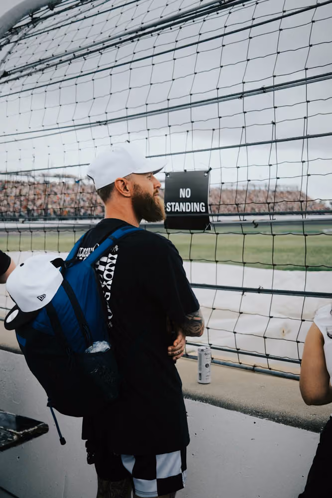 Bearded man wearing white cap and backpack stands near fence with a 'No Standing' sign at sports event.