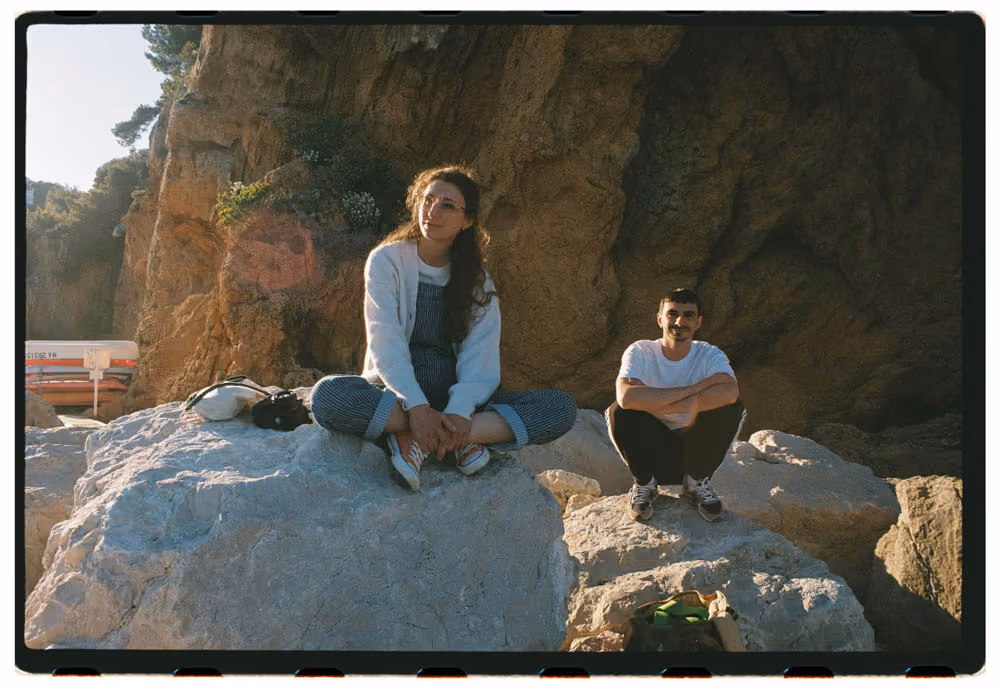 A woman and a man sitting on large rocks near a rocky cliff in the sunlight.