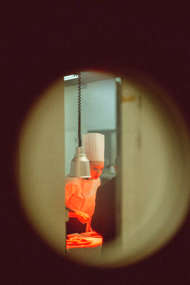 Chef wearing a white hat and uniform preparing food under a heat lamp in a kitchen.