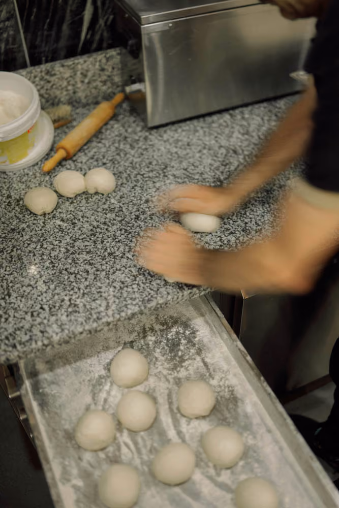 Person shaping dough balls on countertop with more dough balls on a flour-dusted tray below.