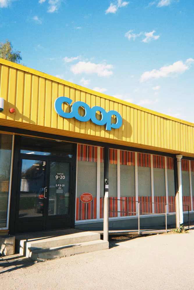 Coop store with yellow facade, glass door showing opening hours, and red-striped window panels.