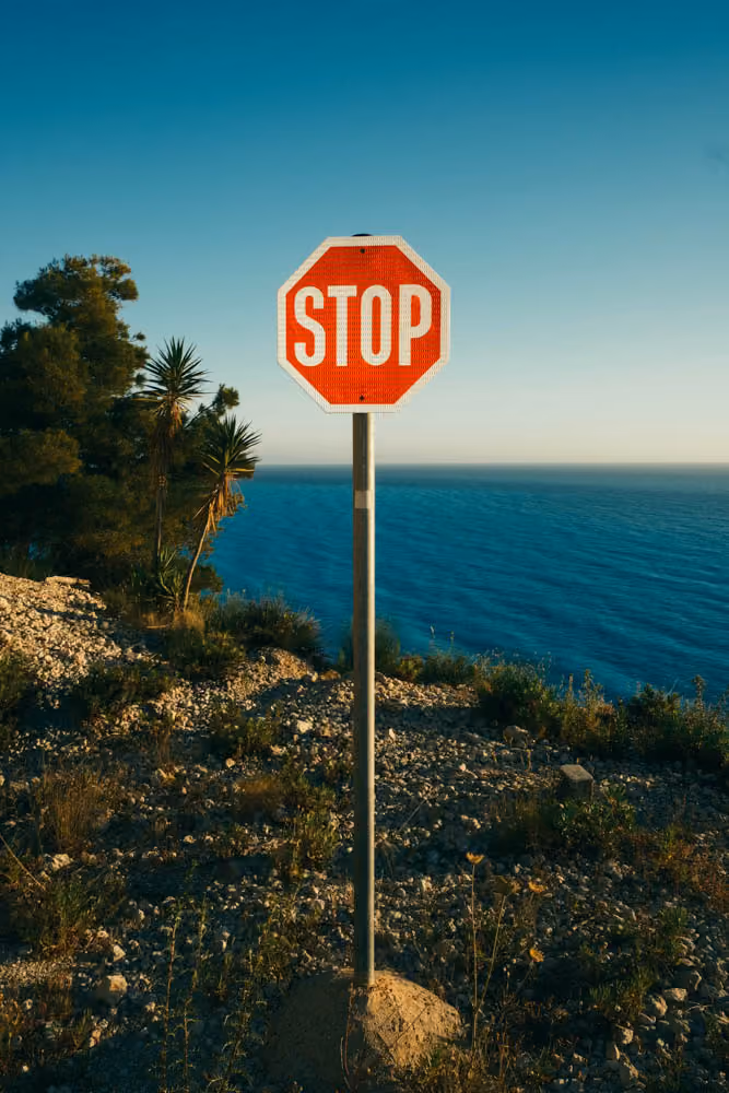 Red stop sign on a pole near rocky coastal terrain with blue ocean and clear sky in background.