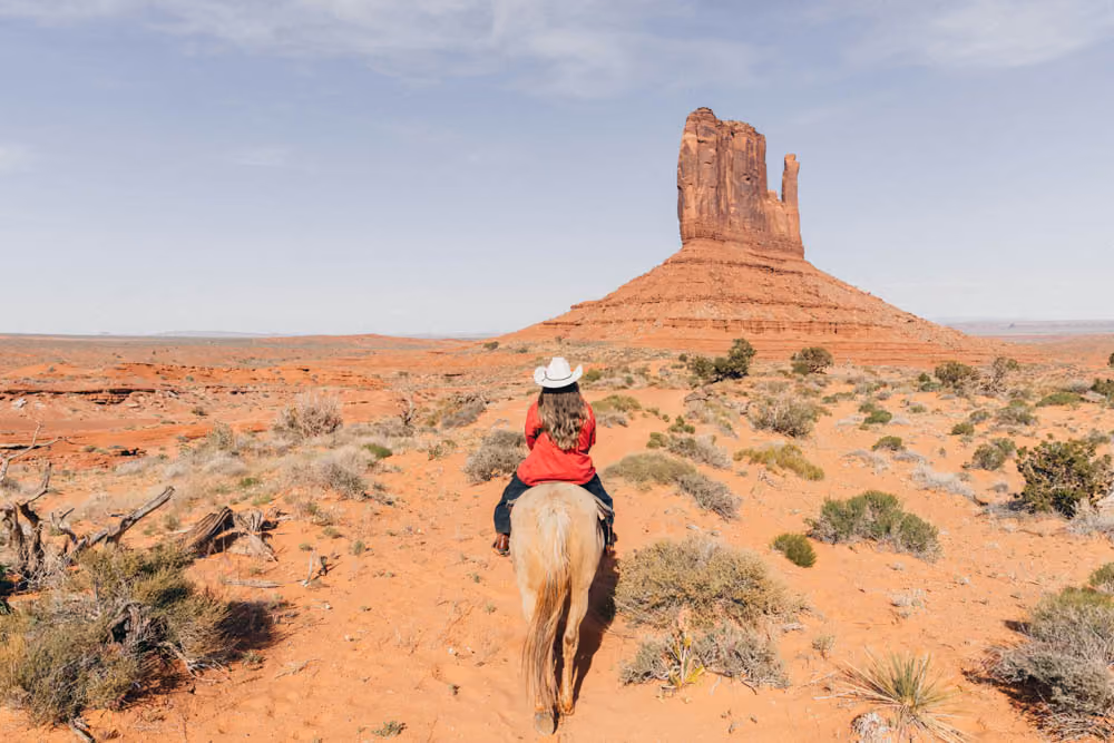 Person in a white hat riding a horse toward a large red rock formation in a desert landscape.