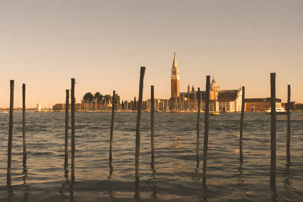 View of Venetian lagoon waters with wooden posts and historic buildings including a tall bell tower at sunset.