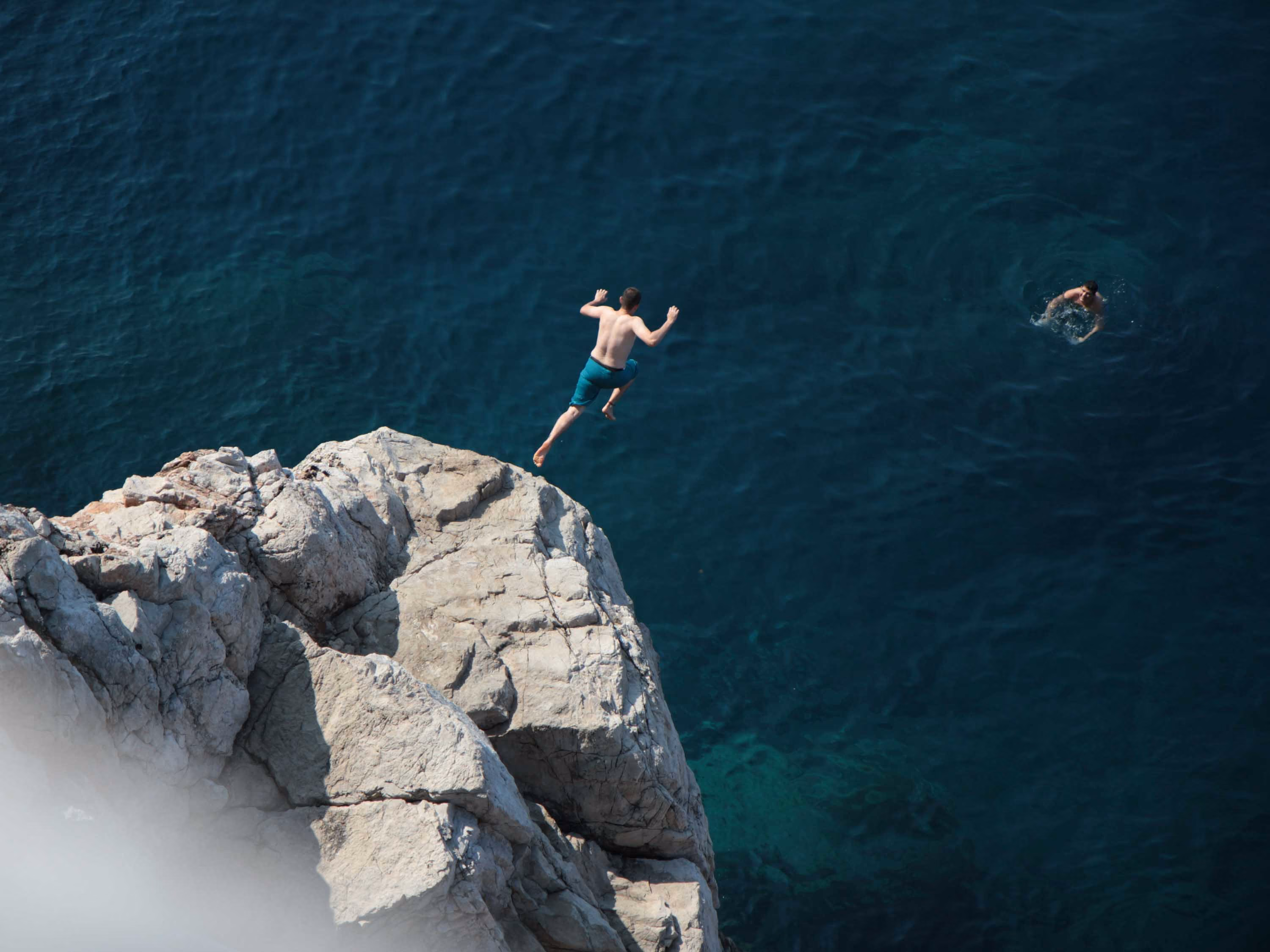 Man mid-air jumping off rocky cliff into dark blue ocean with another swimmer nearby.