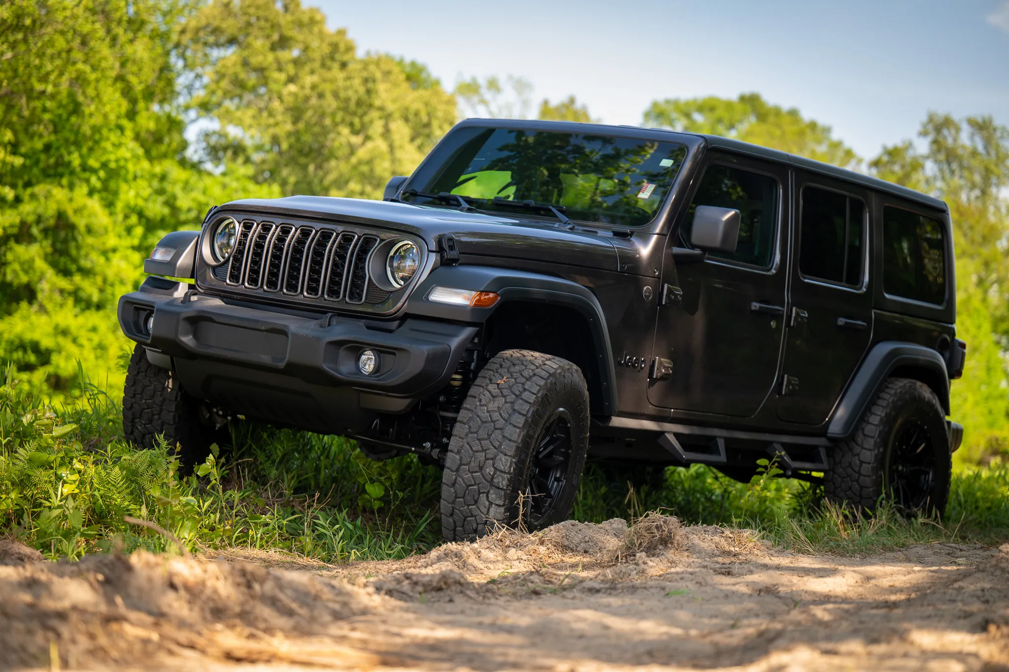 Dark gray Jeep Wrangler Unlimited with black off-road wheels and aggressive all-terrain tires, parked on a dirt trail surrounded by green trees