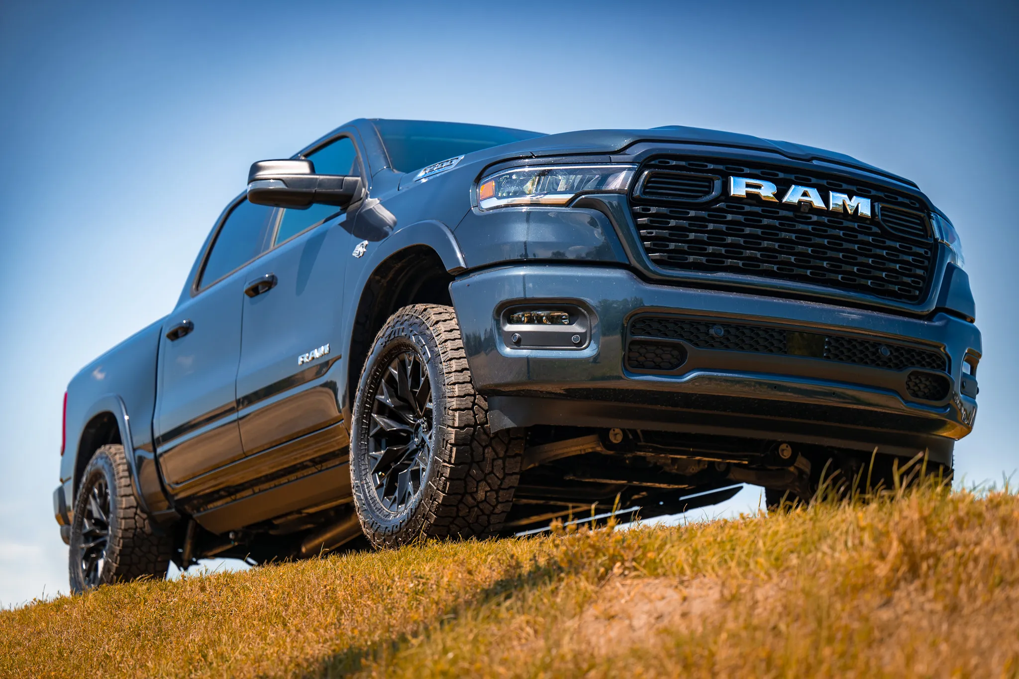 Dark blue Ram 1500 crew cab with black aftermarket wheels and all-terrain tires, photographed from a low angle on a grassy hillside against a clear blue sky