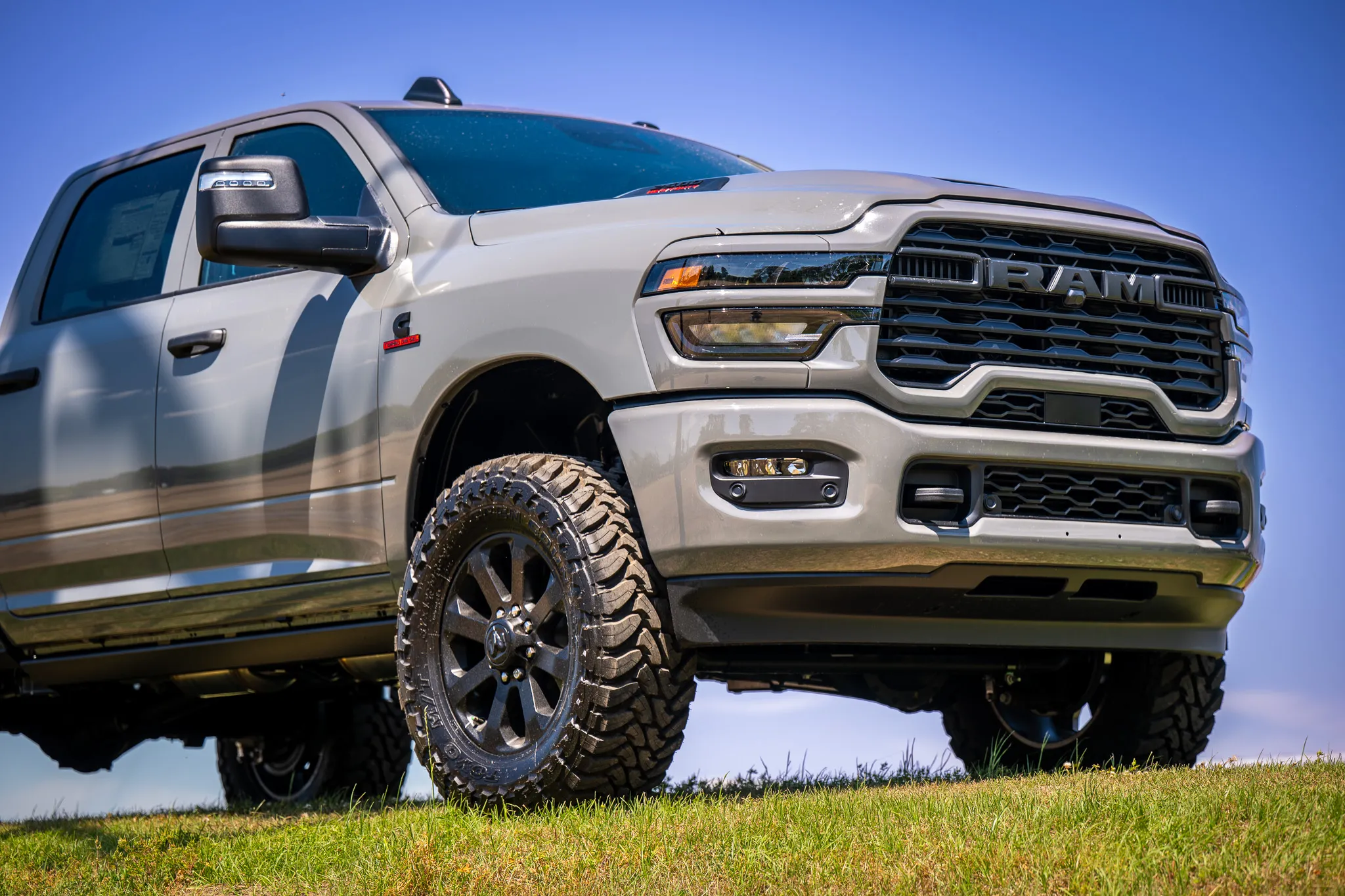low-angle view of a silver ram 2500 heavy duty with dark off-road wheels and mud-terrain tires, parked on a grassy hill against a clear blue sky