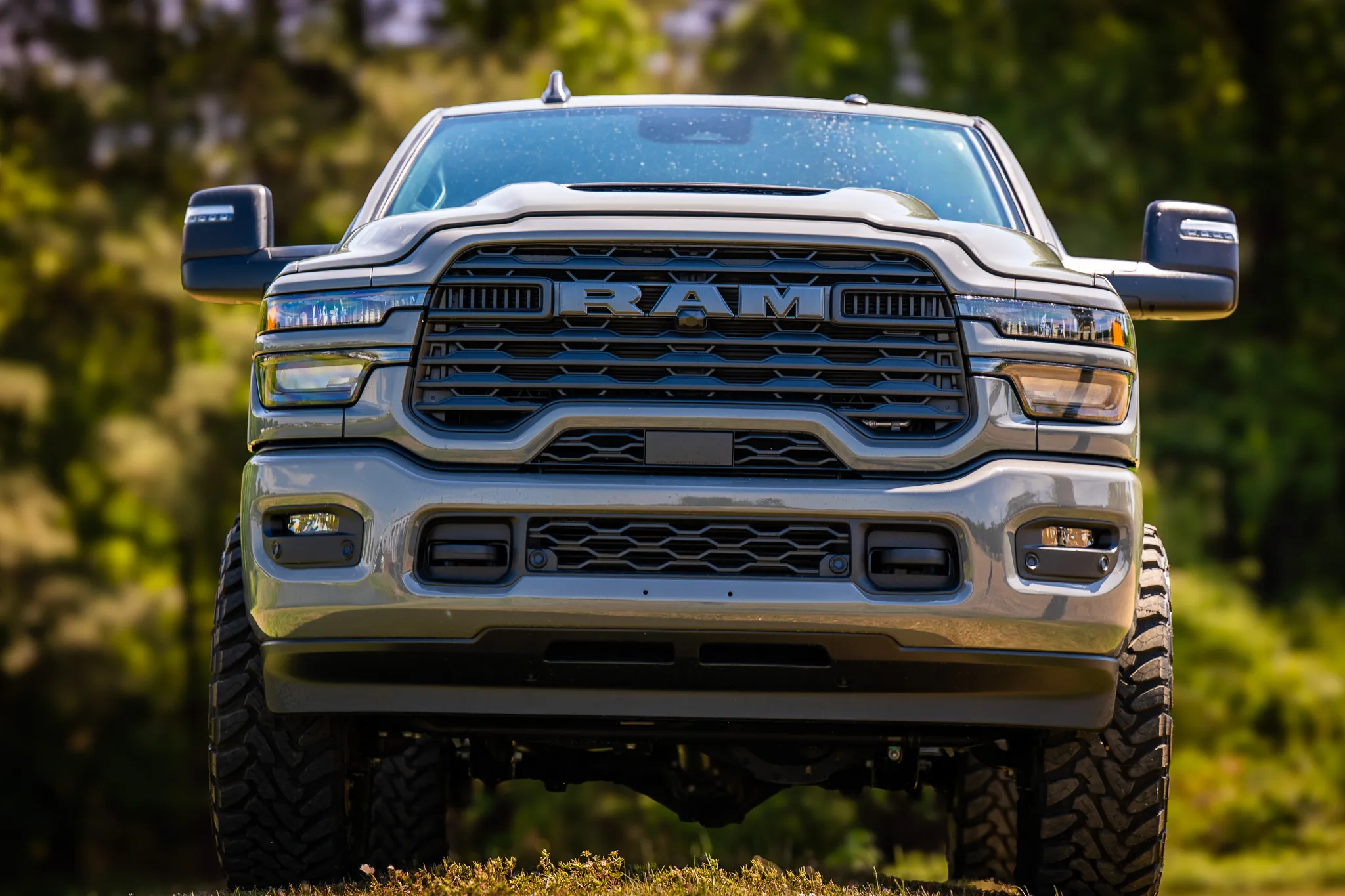  head-on front view of a lifted silver ram 2500 with tow mirrors and mud-terrain tires in warm golden hour light with green trees behind