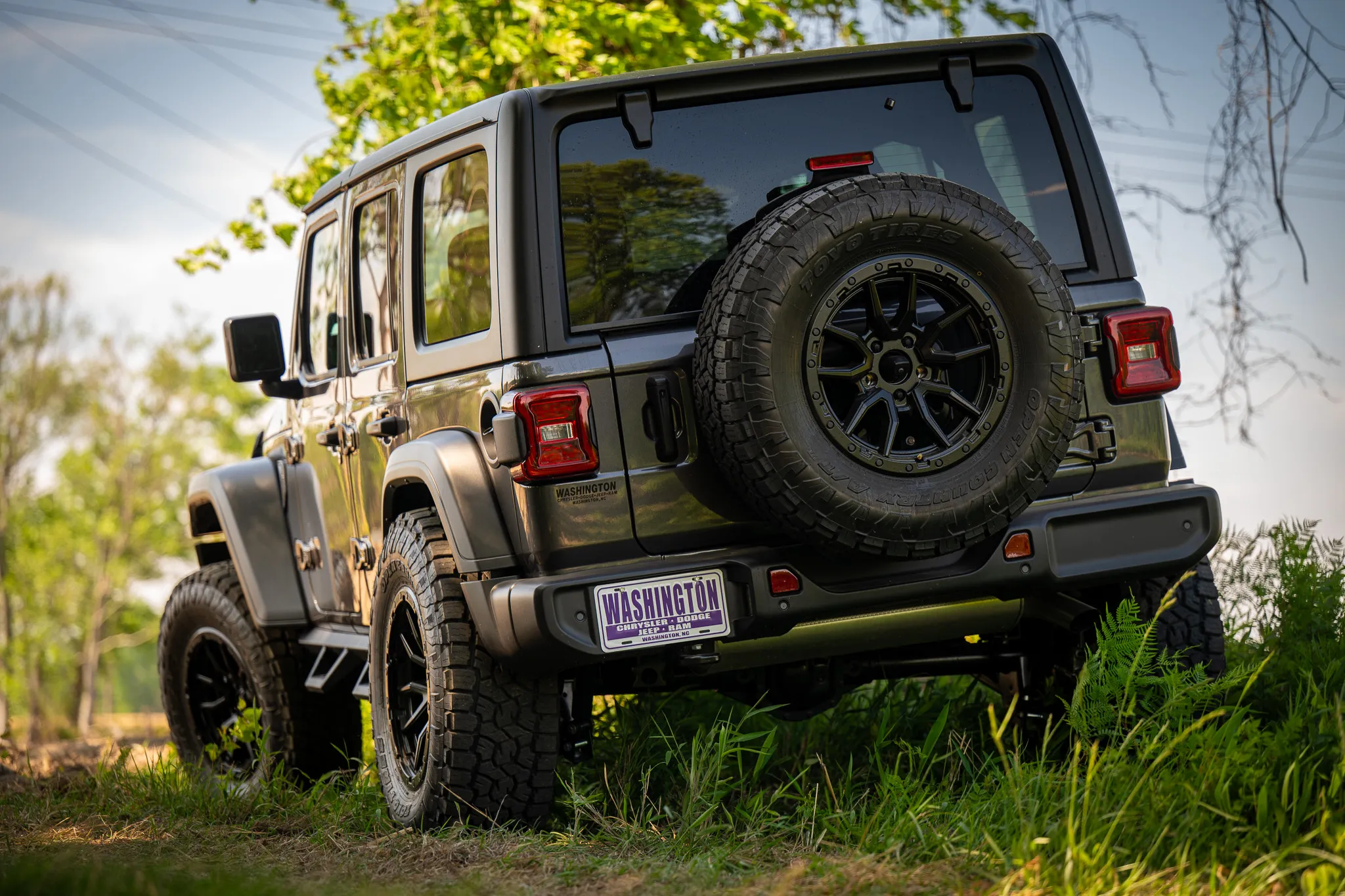 rear three-quarter view of a dark gray jeep wrangler unlimited with black off-road wheels, toyo all-terrain tires, and a matching spare, parked on a grassy trail