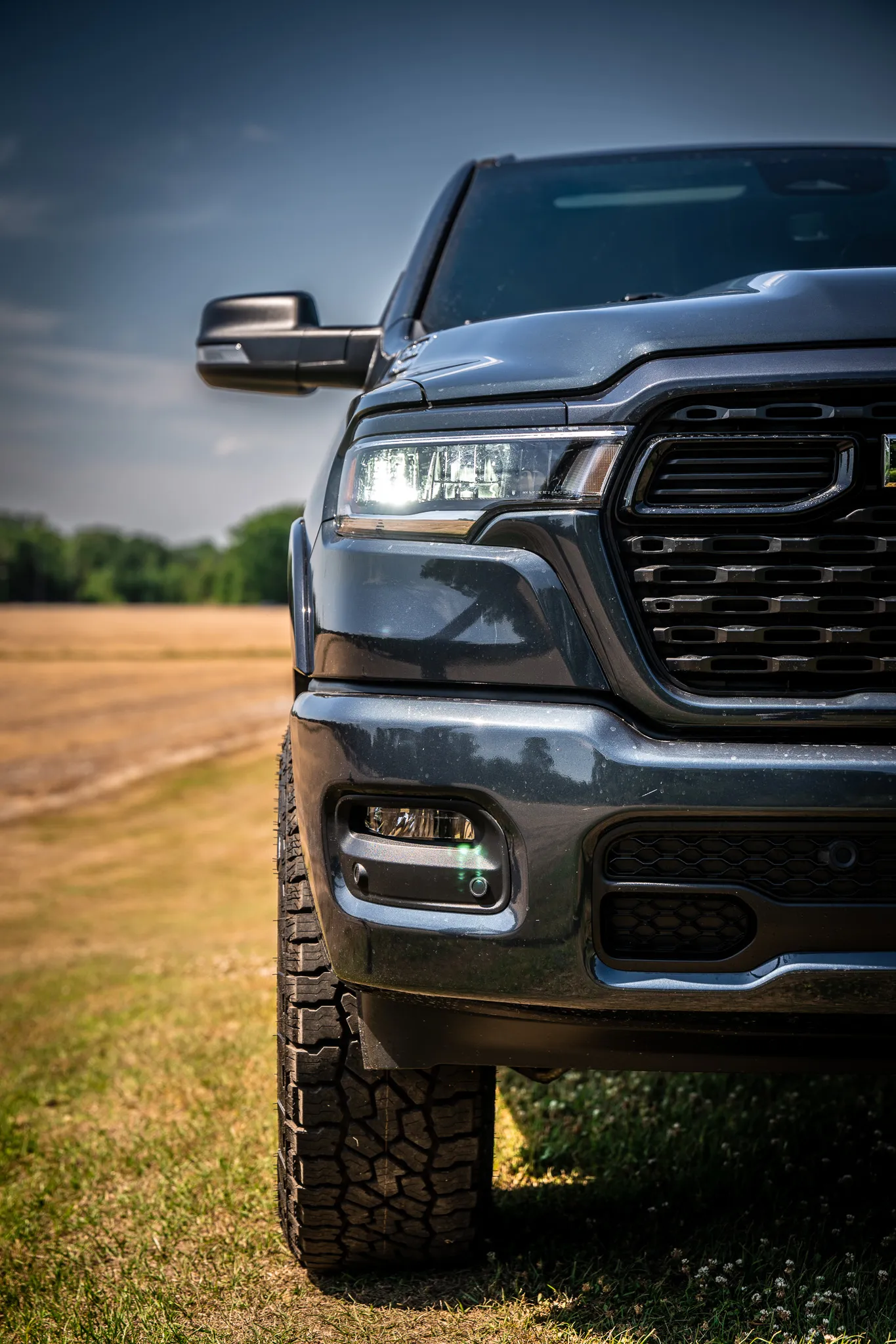 Close-up of a dark blue Ram 1500 front end showing the headlight, grille, and aggressive all-terrain tire tread, parked in an open field