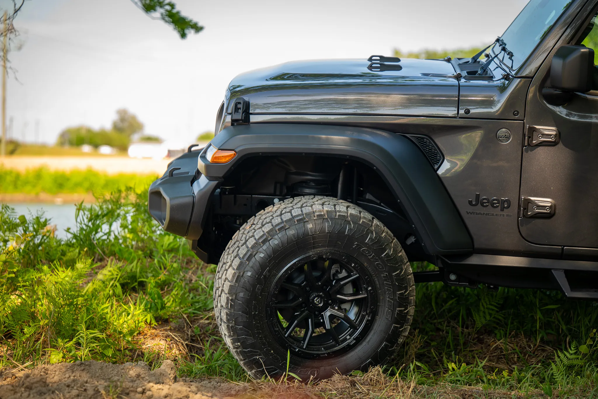 close-up of a dark gray jeep wrangler front wheel showing a black off-road wheel with toyo all-terrain tire and the wrangler badge on the fender