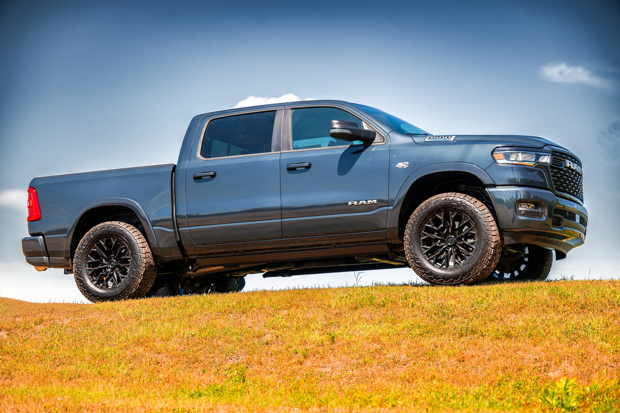 Full side profile of a dark blue ram 1500 crew cab with black aftermarket wheels and all-terrain tires, positioned on a grassy ridge against a blue sky