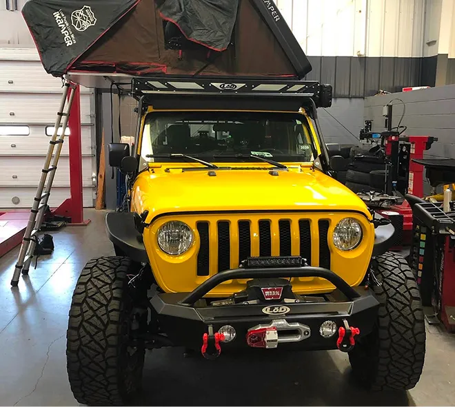 yellow jeep wrangler in a service bay with an ikamper rooftop tent, lod front bumper, warn winch, led light bar, red recovery shackles, and off-road tires