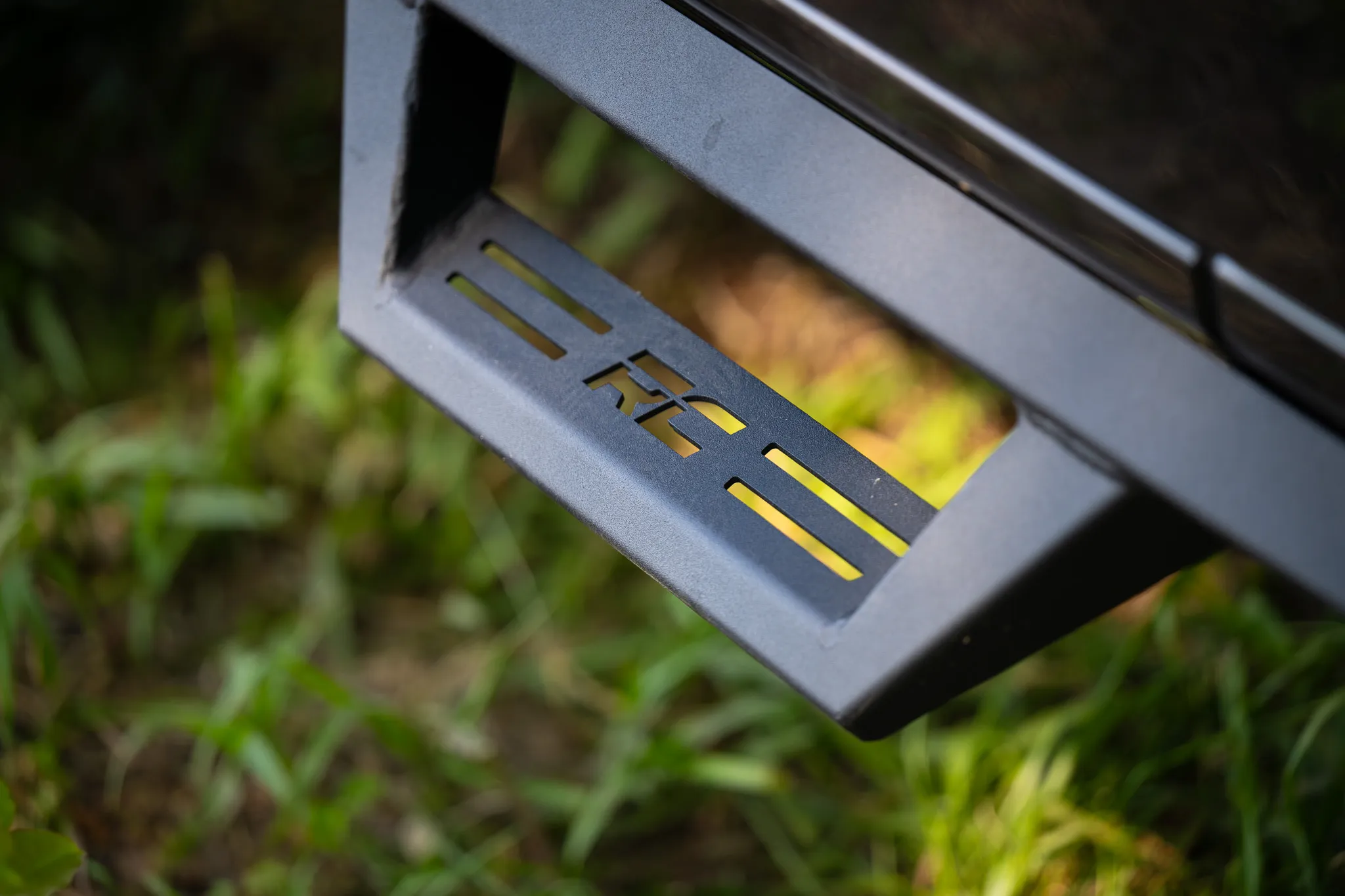 Close-up of a black steel rough country rock slider with laser-cut logo, mounted on a vehicle above green grass