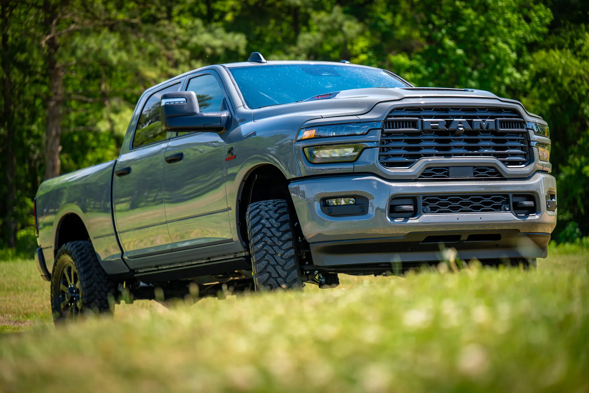 front three-quarter view of a silver ram 2500 cummins with black wheels, mud-terrain tires, and tow mirrors parked on a grassy field with trees behind