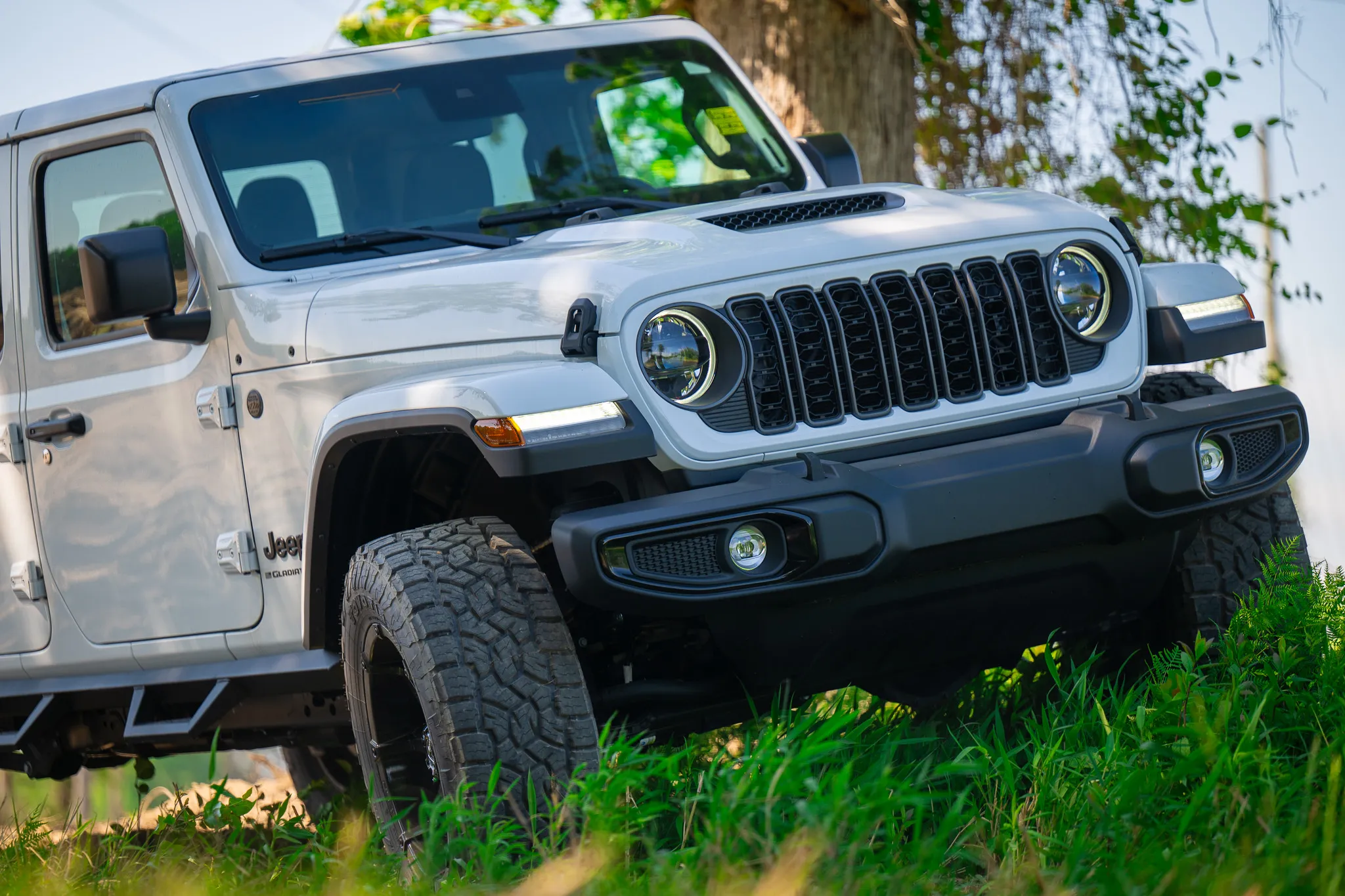 low-angle front quarter view of a white jeep gladiator with off-road tires, rock sliders, and halo led headlights in tall green grass
