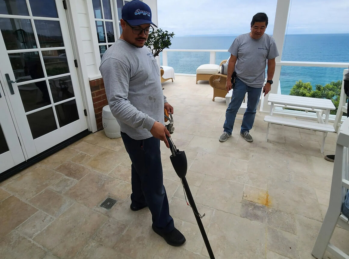 Two men on a tiled patio overlooking the ocean, one holding a metal detector scanning the ground.