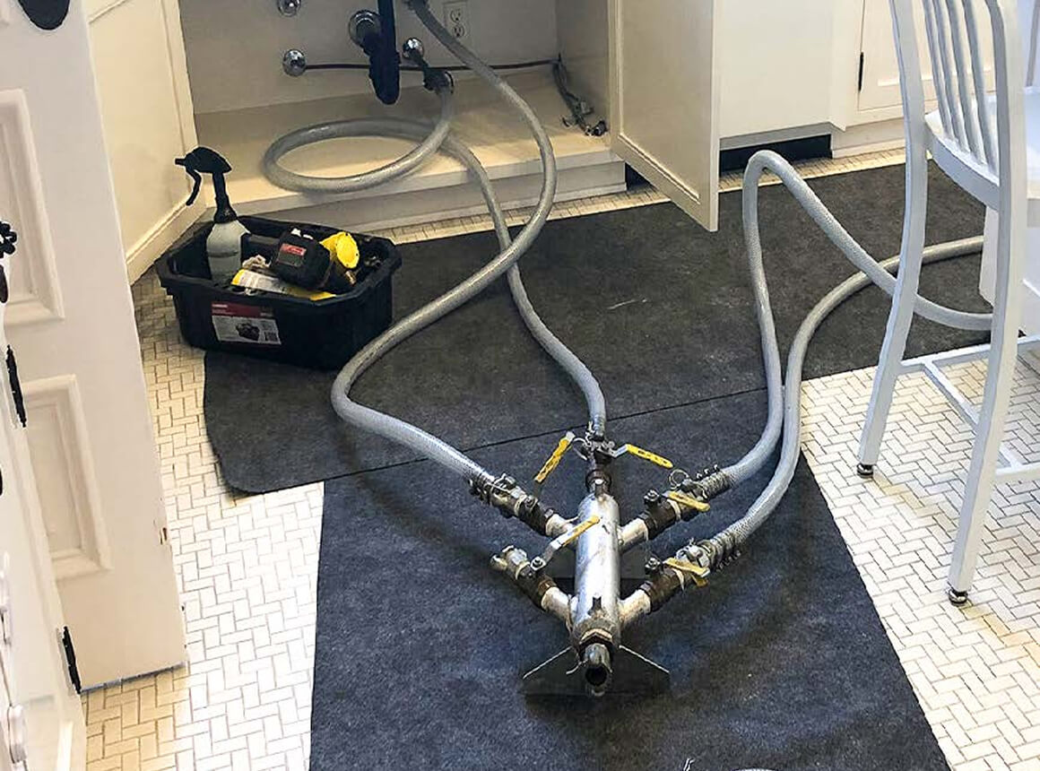 Plumbing tools and hoses connected to valves lying on a dark mat on a tiled kitchen floor with an open cabinet door.