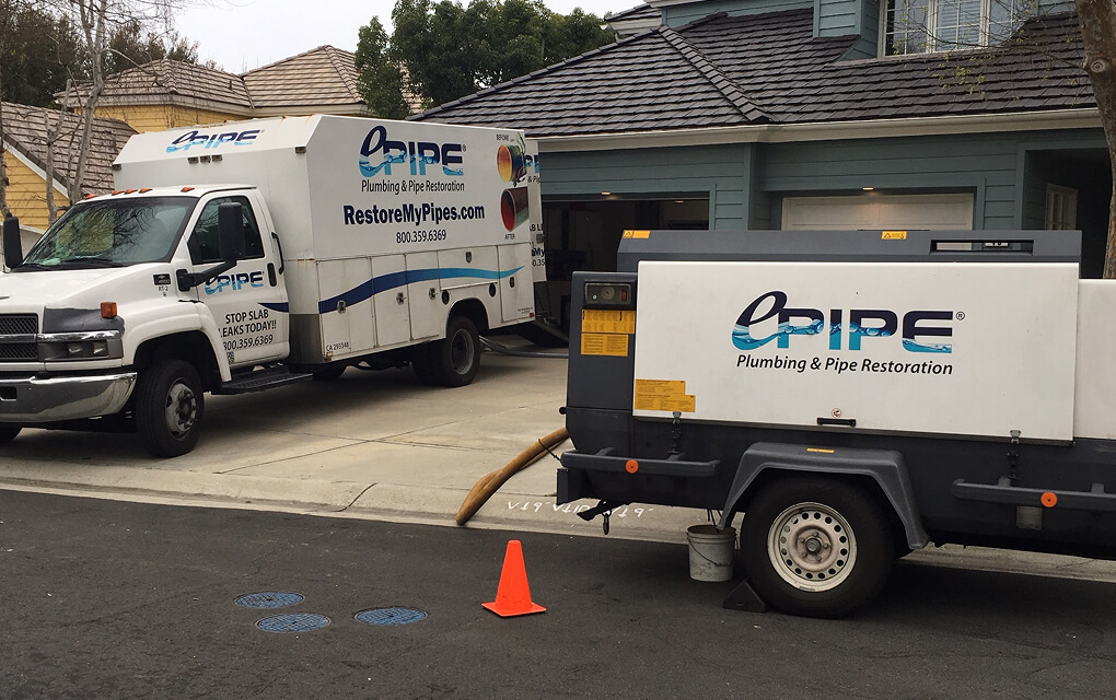 Two ePIPE Plumbing & Pipe Restoration vehicles parked outside a residential driveway with a hose connected to the house and an orange traffic cone on the street.