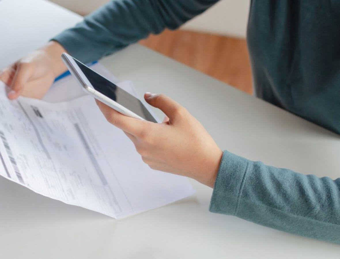 Person holding a smartphone in one hand and a document in the other at a white desk.