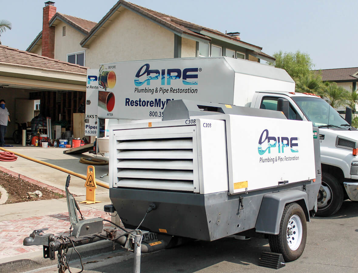 Worksite with a white plumbing restoration truck and equipment trailer parked in front of a house under renovation.