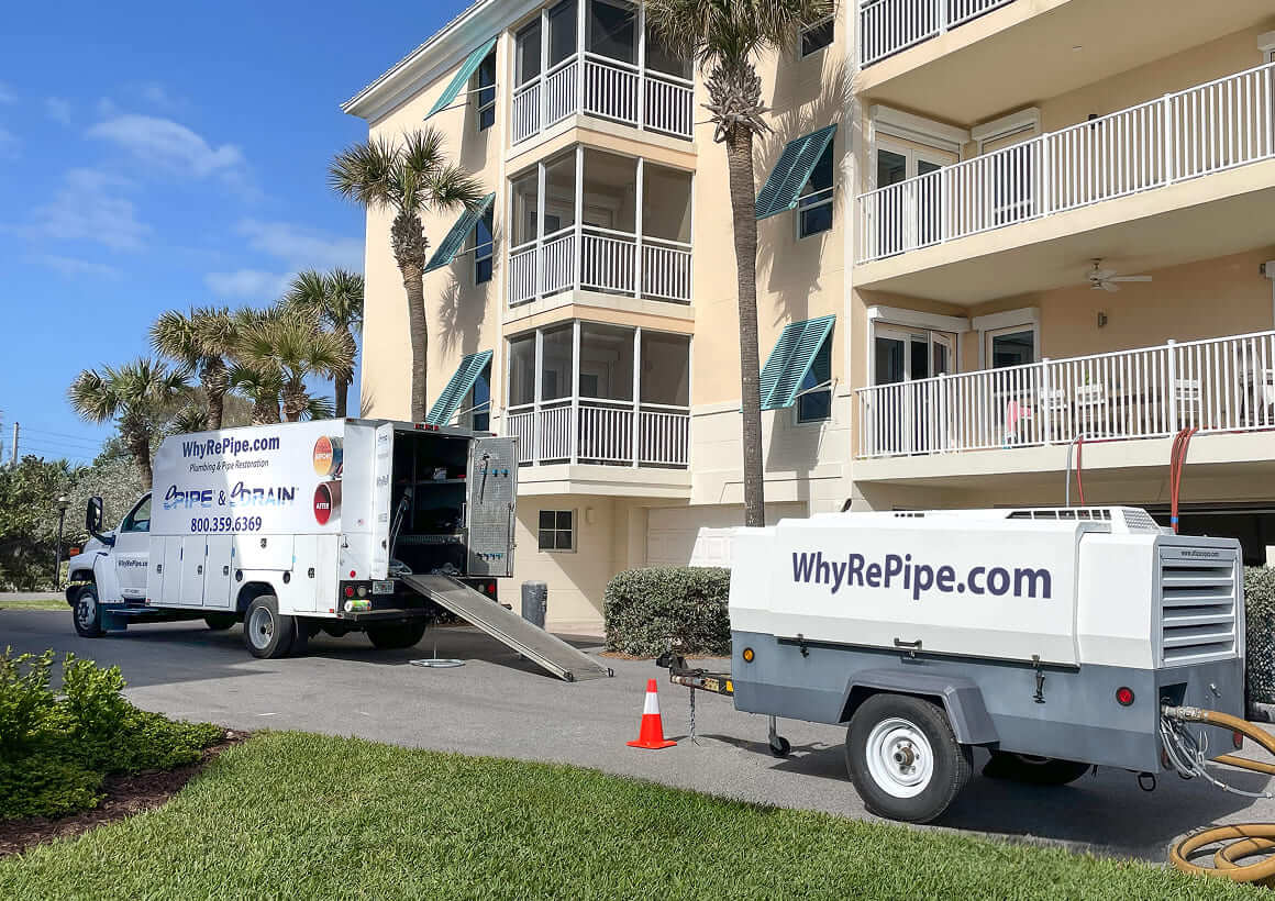 Service truck and equipment trailer from WhyRePipe.com parked outside an apartment building with palm trees and green grass.
