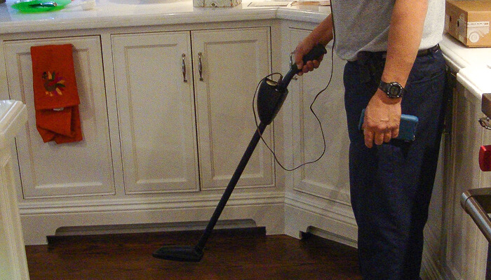 Person using a handheld vacuum cleaner to clean the floor in a kitchen with white cabinets and dark wood flooring.