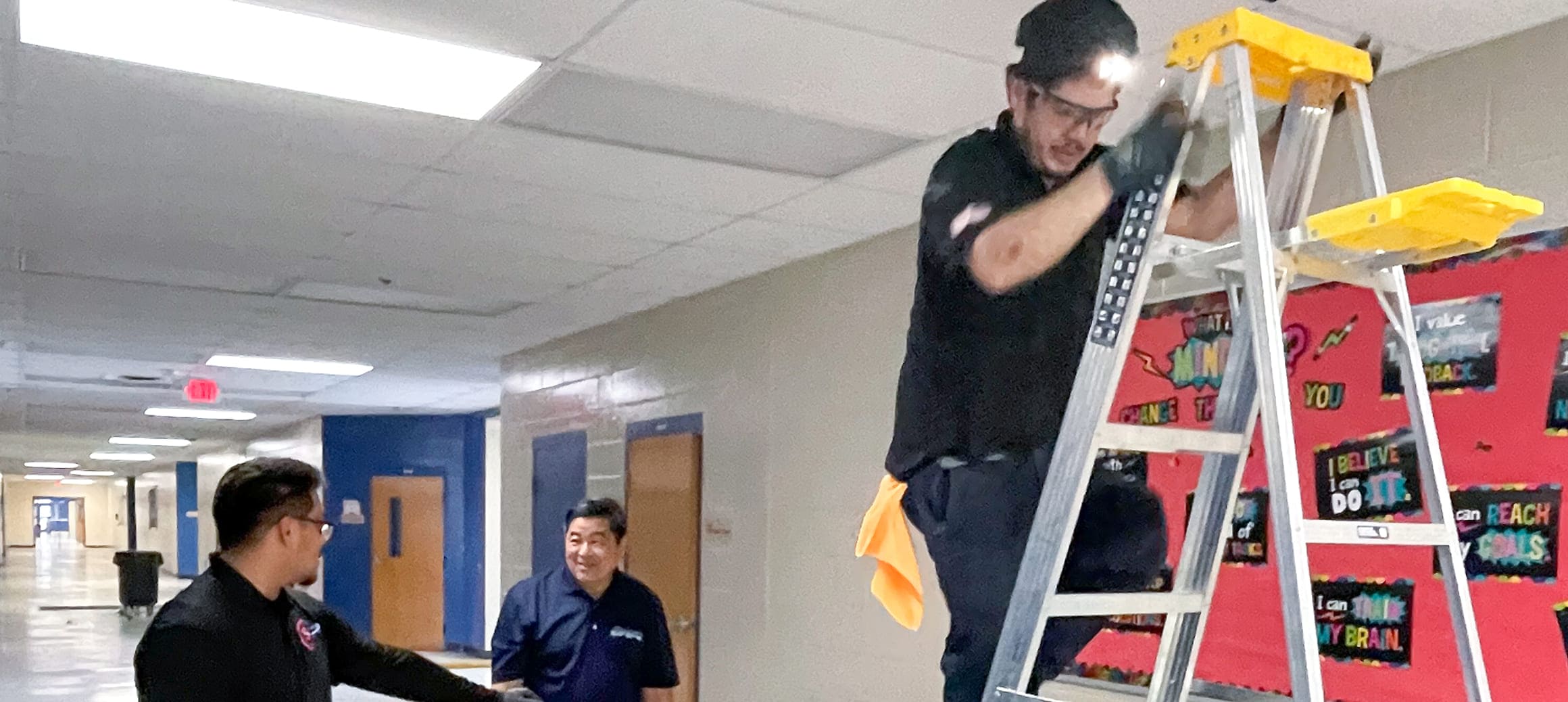 A technician wearing a headlamp and safety glasses standing on a ladder in a hallway, another person pointing and a third person watching nearby.