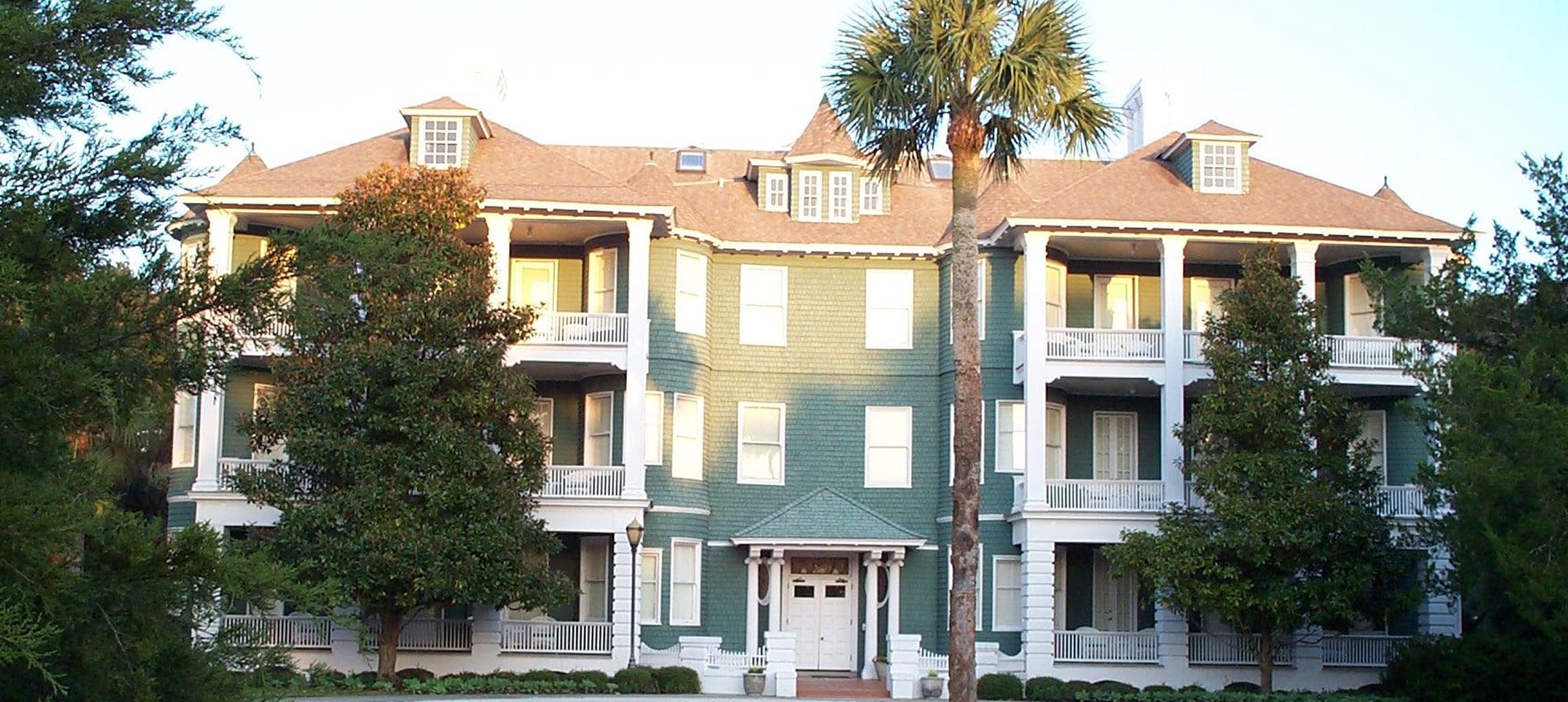Large green multi-story house with white trim, balconies, and surrounded by trees including a prominent palm tree.
