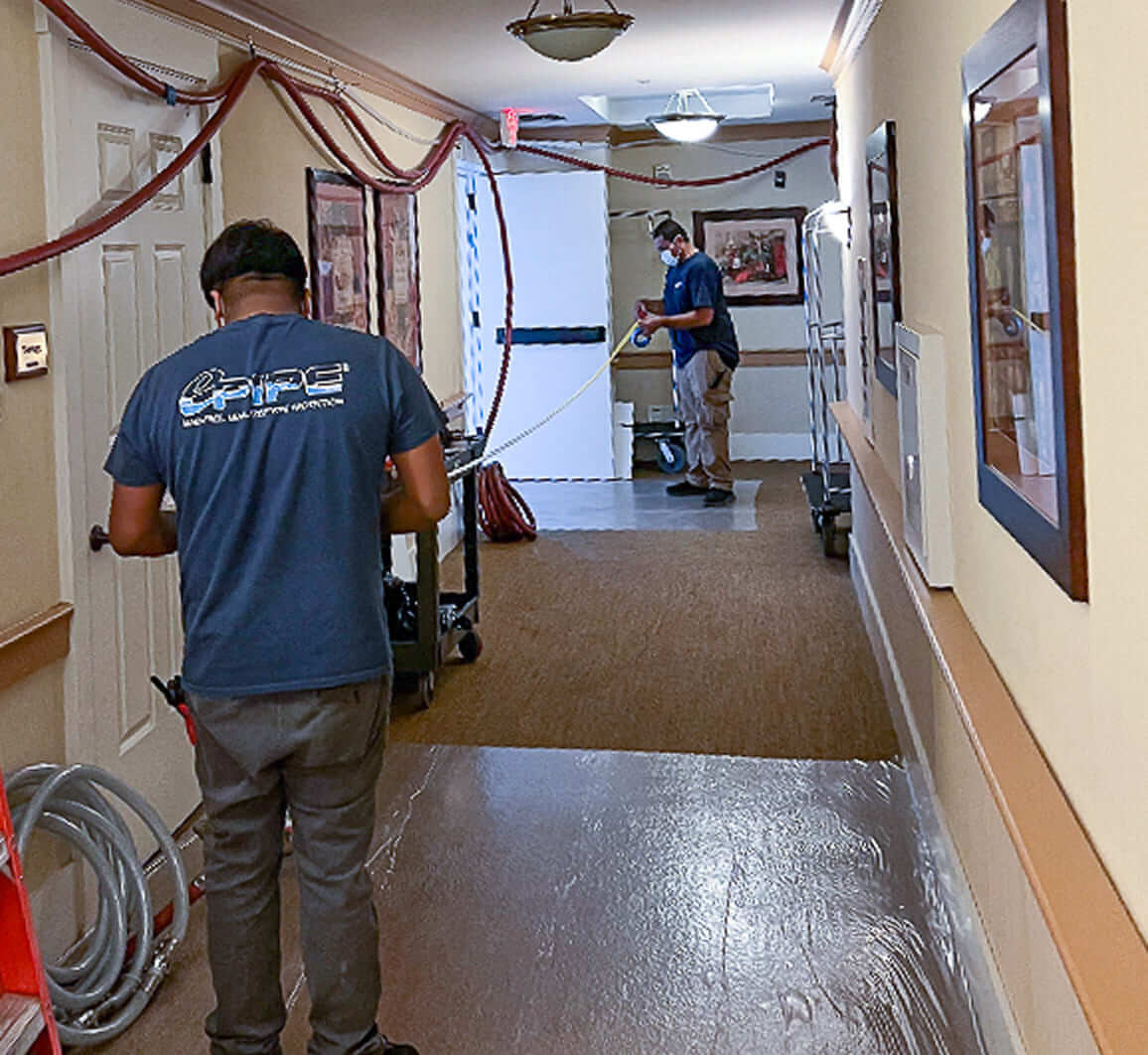 Two workers in a hallway installing or inspecting large hoses, with one worker wearing a mask and stretching a yellow tape measure.