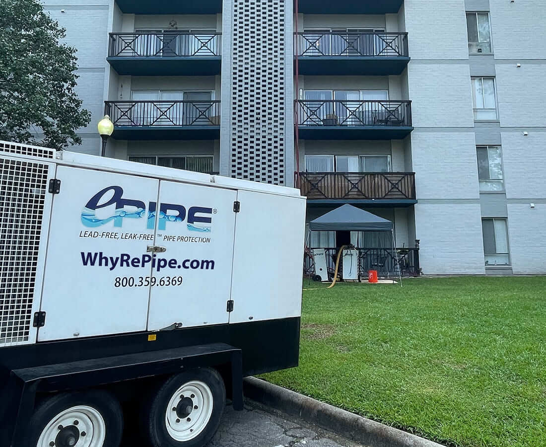 Outdoor setup in front of an apartment building with a white trailer labeled 'O-Pipe Lead-Free, Leak-Free Pipe Protection' and website WhyRePipe.com visible.