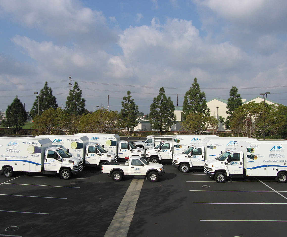 Fleet of white service trucks and a pickup parked in a lot with trees and buildings in the background under a cloudy sky.