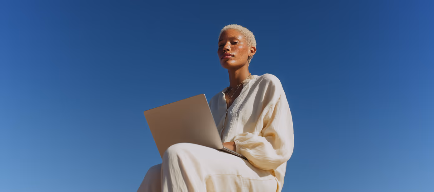 Person with short blonde hair wearing cream clothing sitting outdoors with a laptop against a clear blue sky.