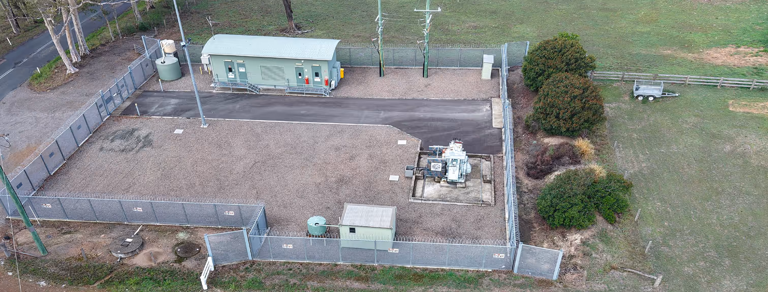 Aerial view of a fenced electrical substation with a control building, transformer, and surrounding gravel yard in a rural setting.