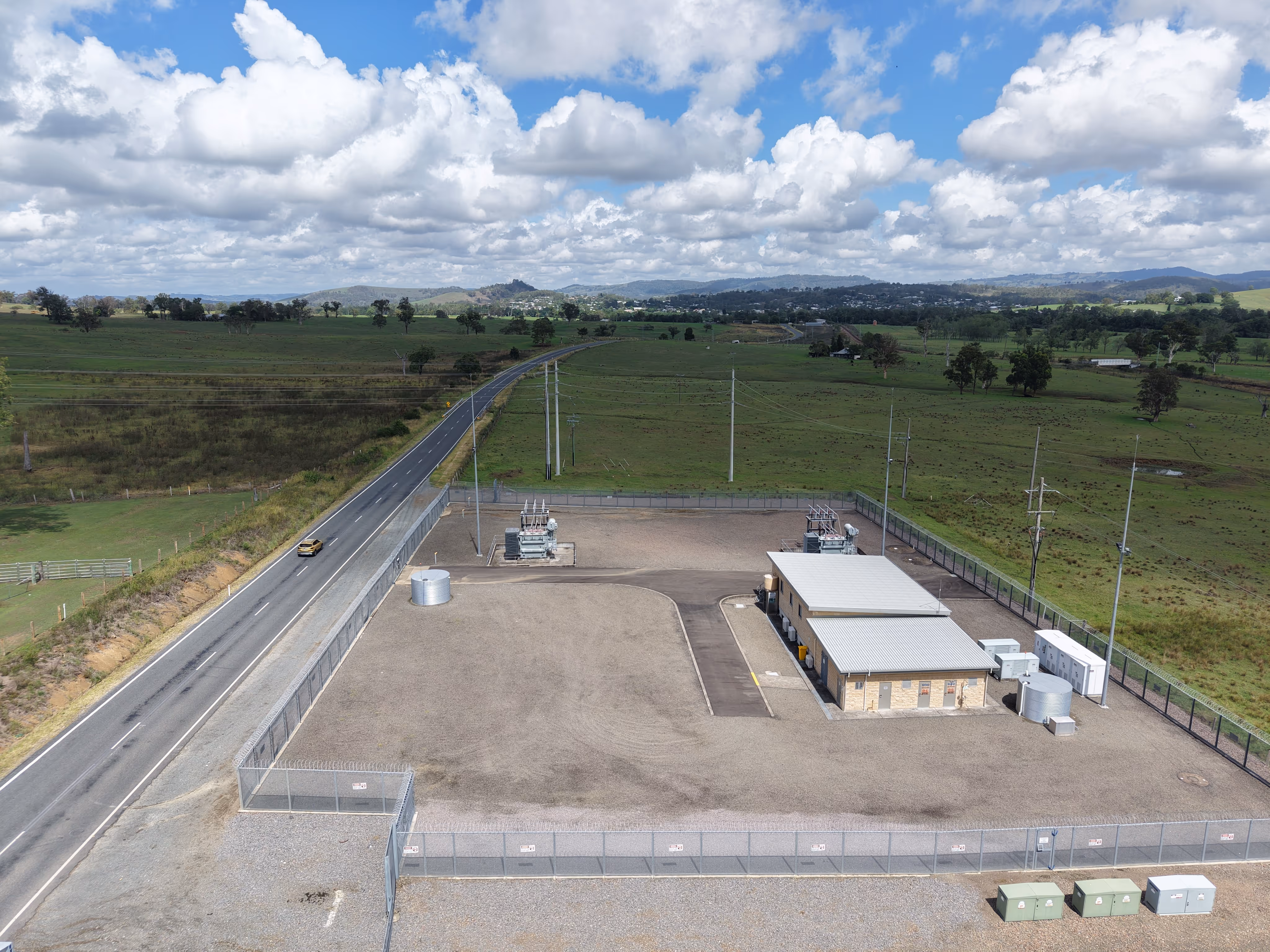 Aerial view of a fenced electrical substation beside a rural highway, with a small switchyard and equipment building on a gravel pad, surrounded by green grazing land and distant hills under a partly cloudy sky.