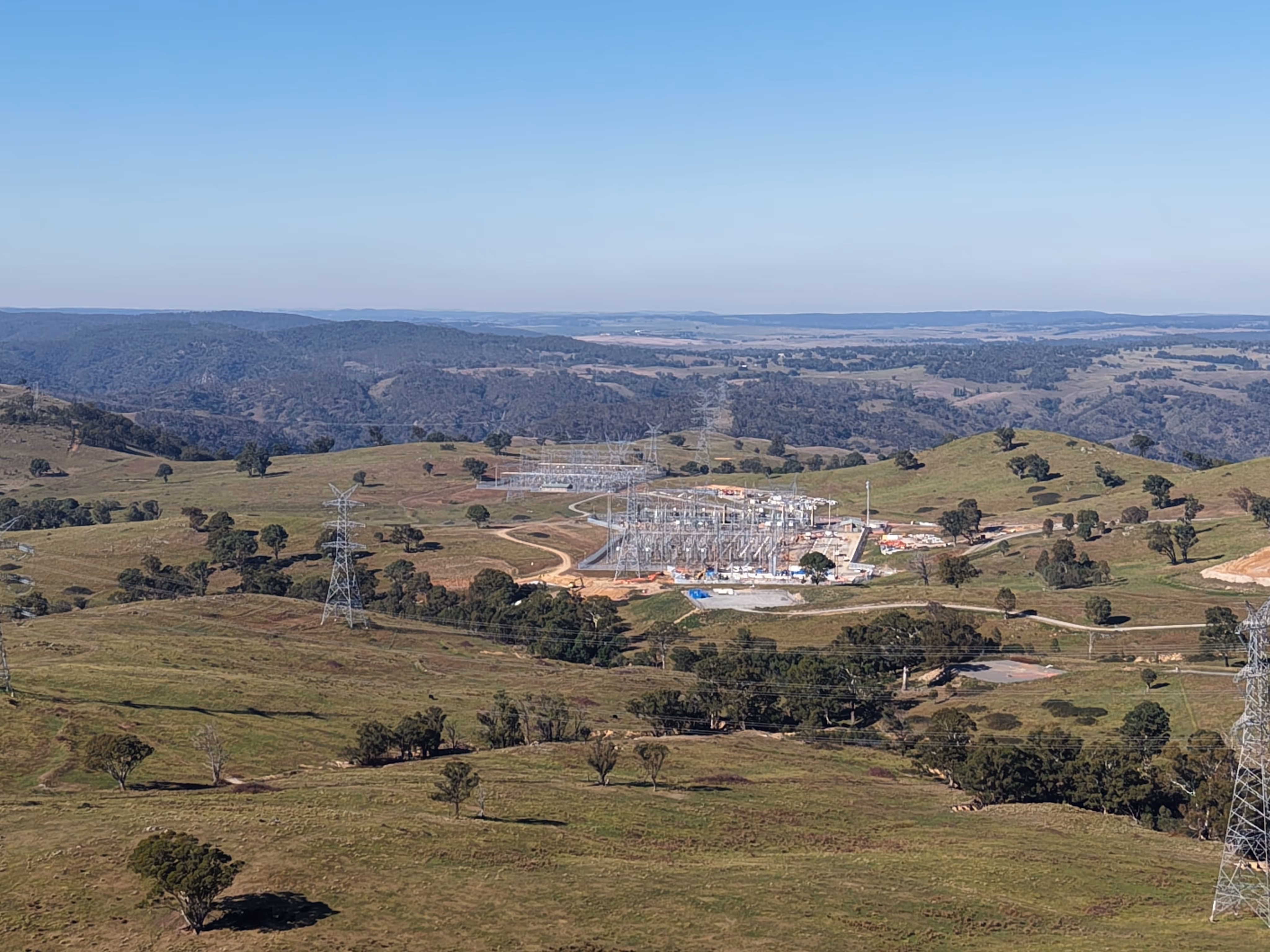 Aerial view of an electrical substation with transmission towers set among rolling pastoral hills and a distant valley.