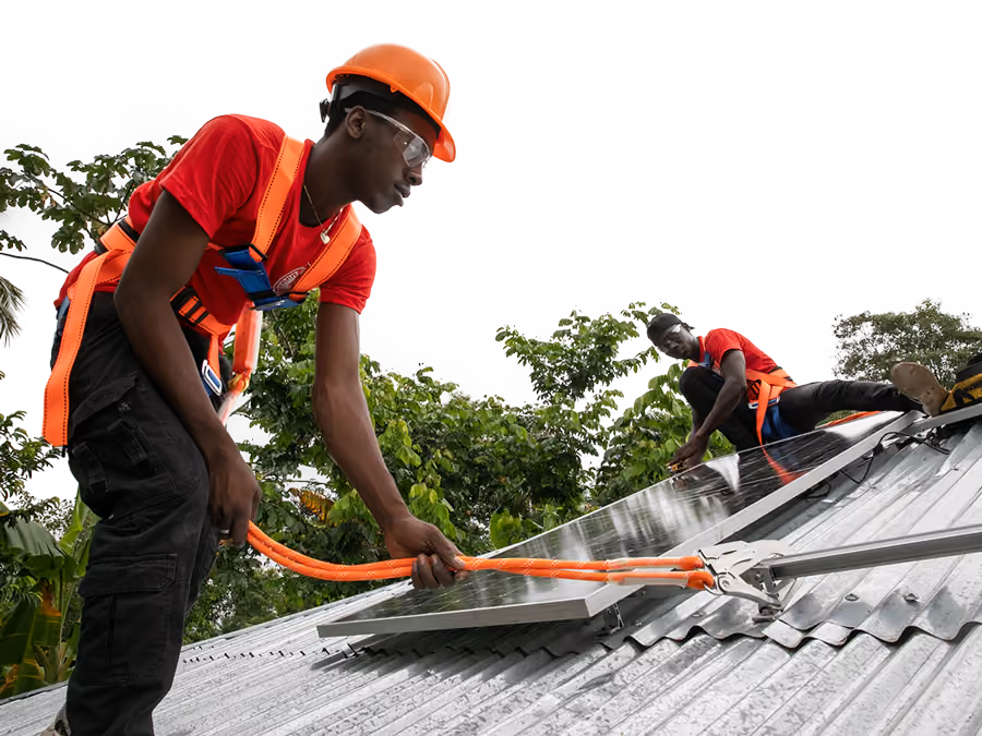 An Alina Eneji installer in a hard hat and safety harness pulls a cable across a metal roof while fitting rooftop solar in Haiti