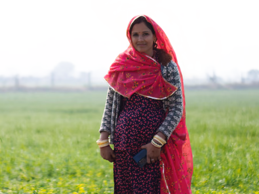 A woman in a pink dupatta holds a mobile phone while standing in a green wheat field in Rajasthan, India