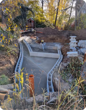 Baustelle mit Betonform für Pool oder Teich, umgeben von Bäumen und Garten mit Messgerät in der Mitte.