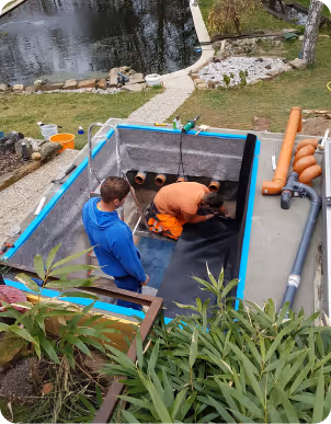 Two men installing a liner inside a rectangular outdoor pond under construction near a garden pond.