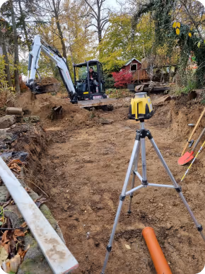 Bagger arbeitet auf einer Baustelle mit Erdbewegungen, davor steht ein Vermessungsgerät auf einem Stativ, umgeben von Bäumen und herbstlicher Vegetation.
