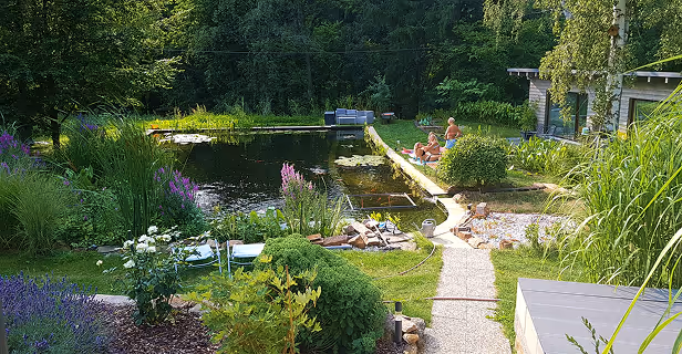 Ein natürlicher Gartenteich mit klaren Wassern, umgeben von Blumen, Sträuchern und Bäumen, mit Menschen, die am Ufer entspannen.
