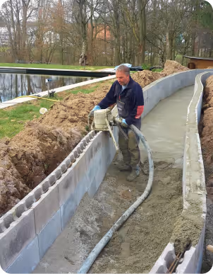 Bauarbeiter glättet geschwungenen Betonweg neben einer Mauer in einer Parklandschaft mit Teich und Bäumen.
