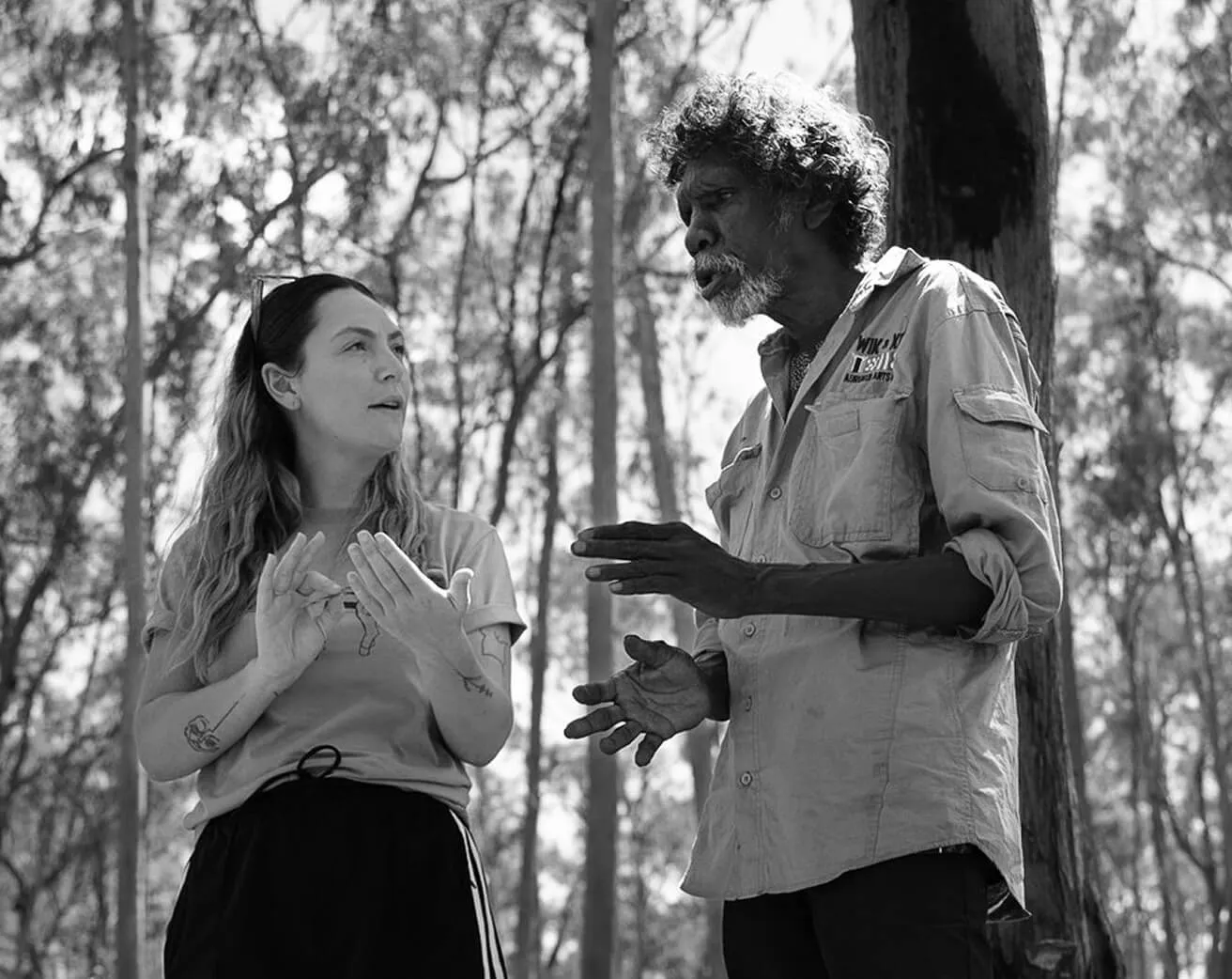 Woman reaching out under the dappled light of trees on Country, black-and-white photograph