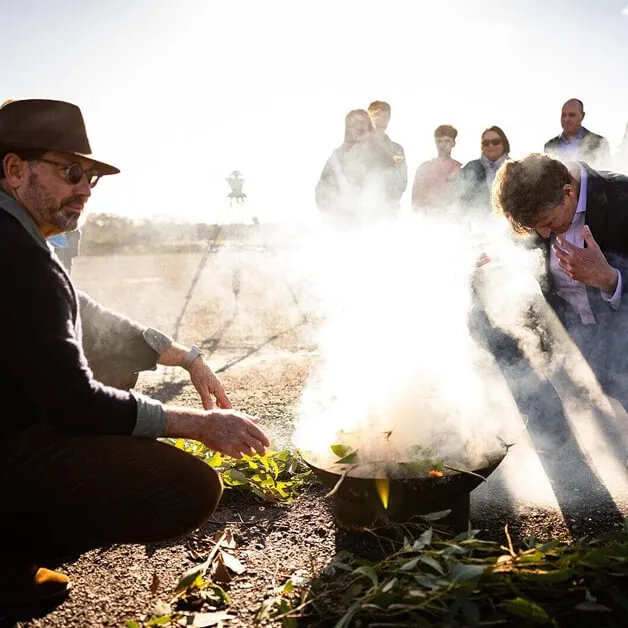 Man in a wide-brimmed hat tending a smoking ceremony over green leaves with community gathered around