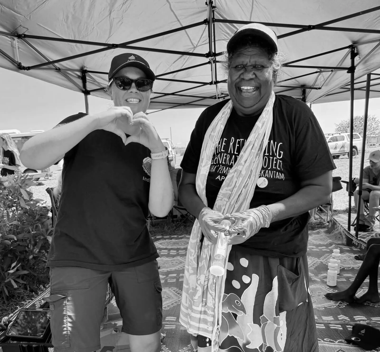 Two men smiling together at a community event under a marquee, one making a heart shape with her hands, black-and-white photograph