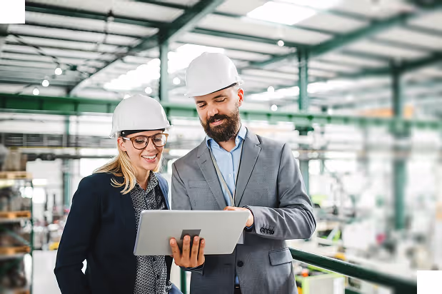 Two engineers in hard hats reviewing information on a tablet inside a large industrial facility.